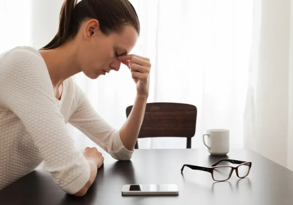 A woman looking stressed while sitting at a table with glasses and a cup.